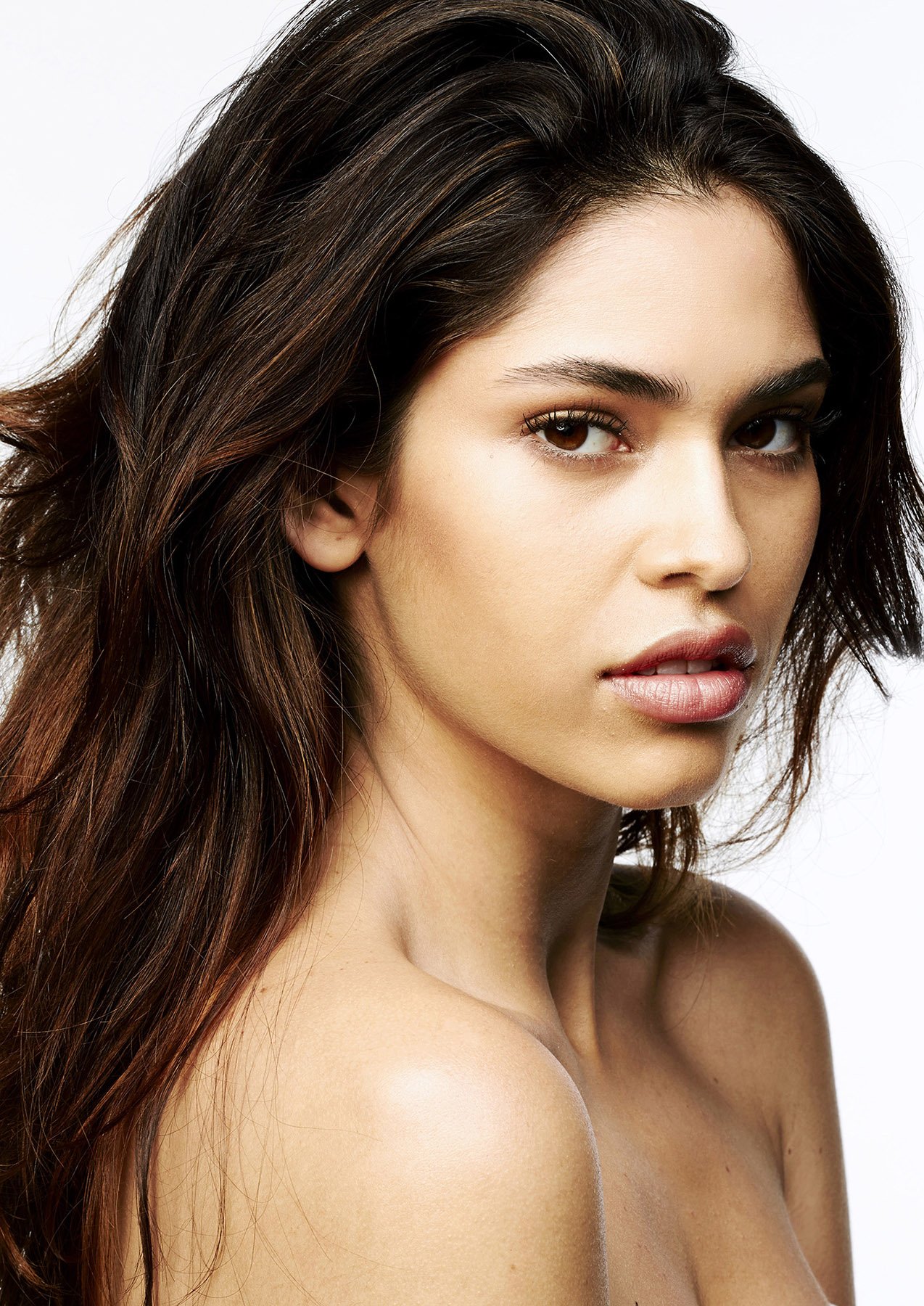 Portrait of a young adult woman with long wavy brown hair and a bare shoulder against a clean white background in a beauty shot.