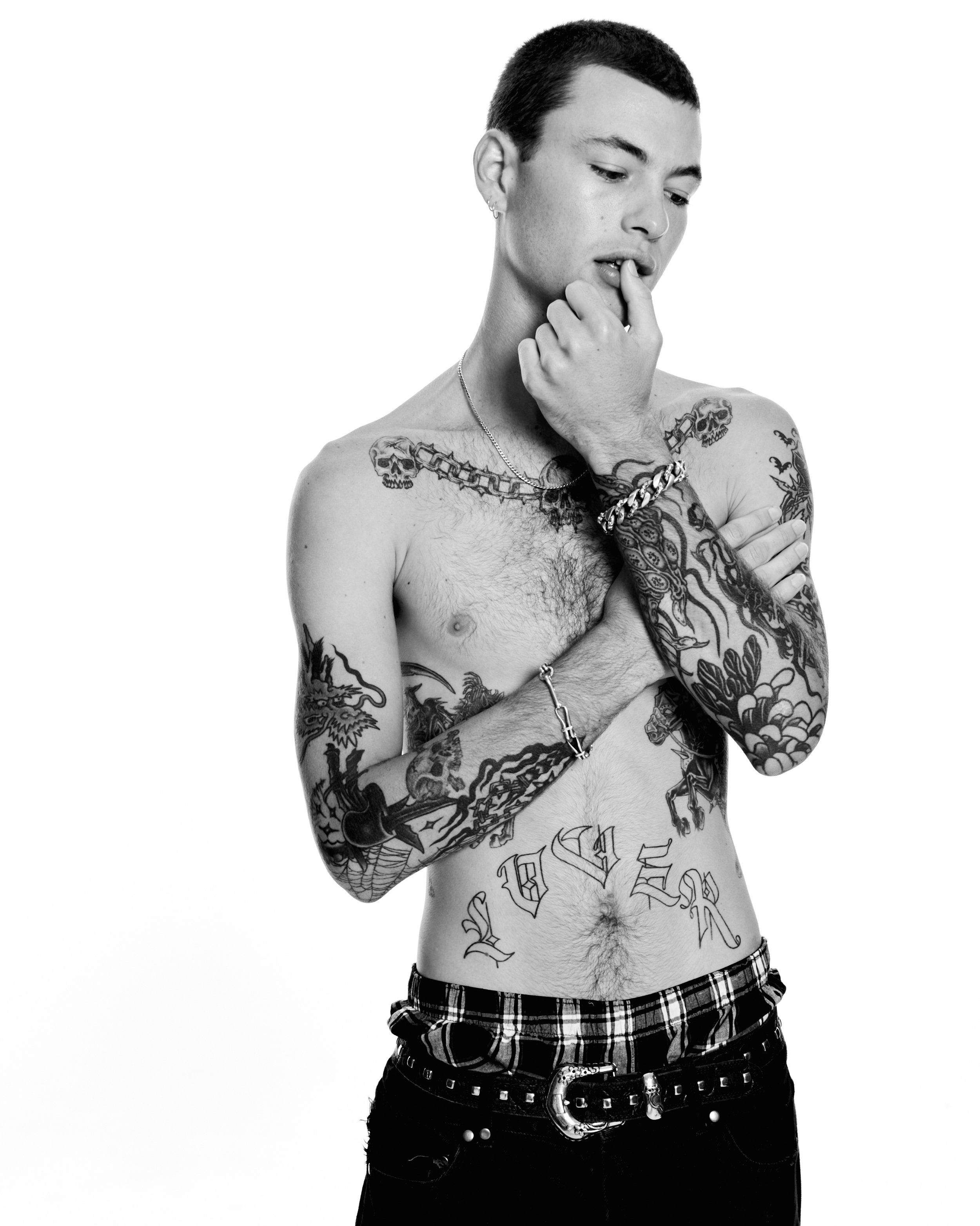Monochrome portrait of a shirtless tattooed man in black pants and a plaid belt, hand near mouth, against a bright white backdrop.