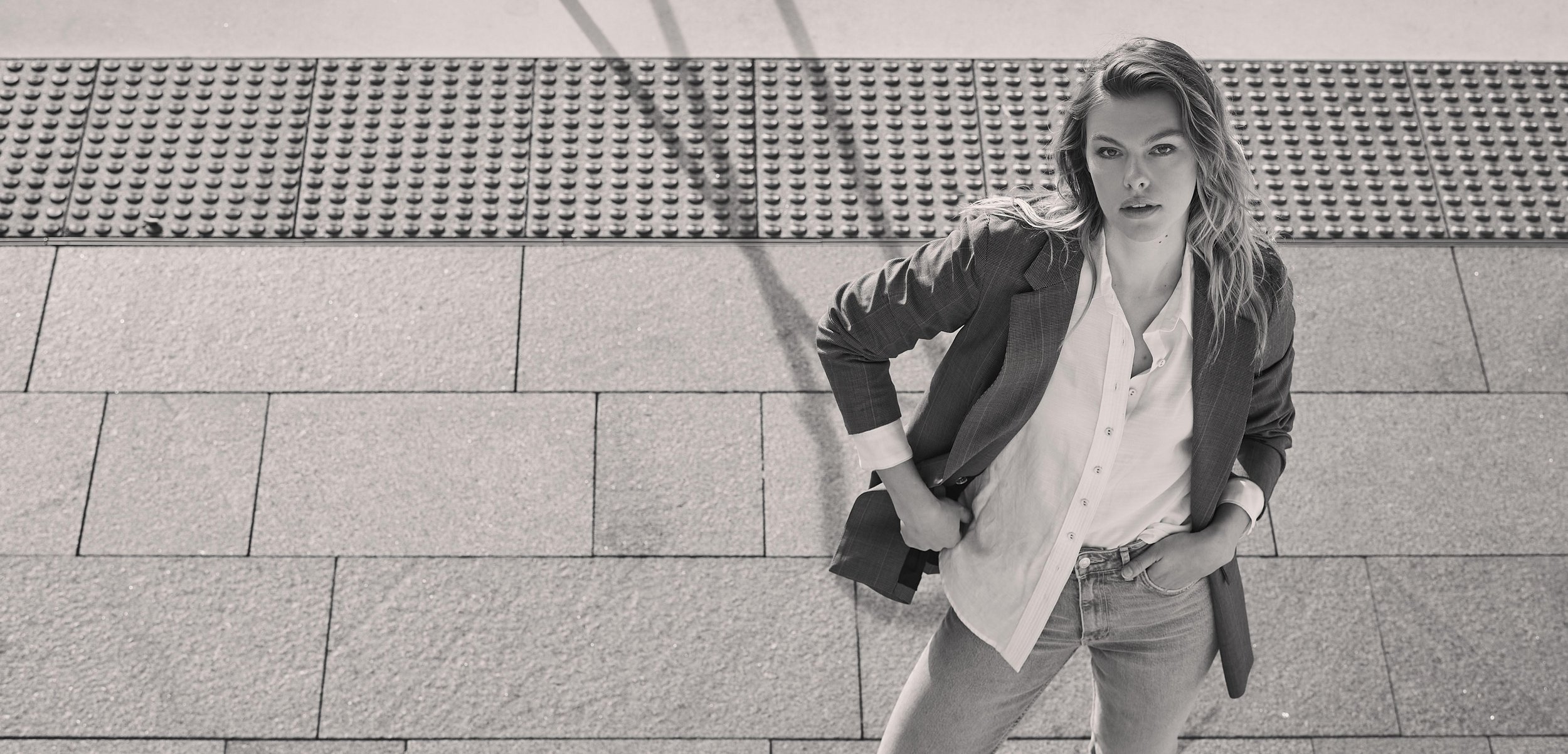 Monochrome medium shot of a stylish adult woman looking at the camera, wearing a blazer, button-up shirt, and jeans against a concrete backdrop.