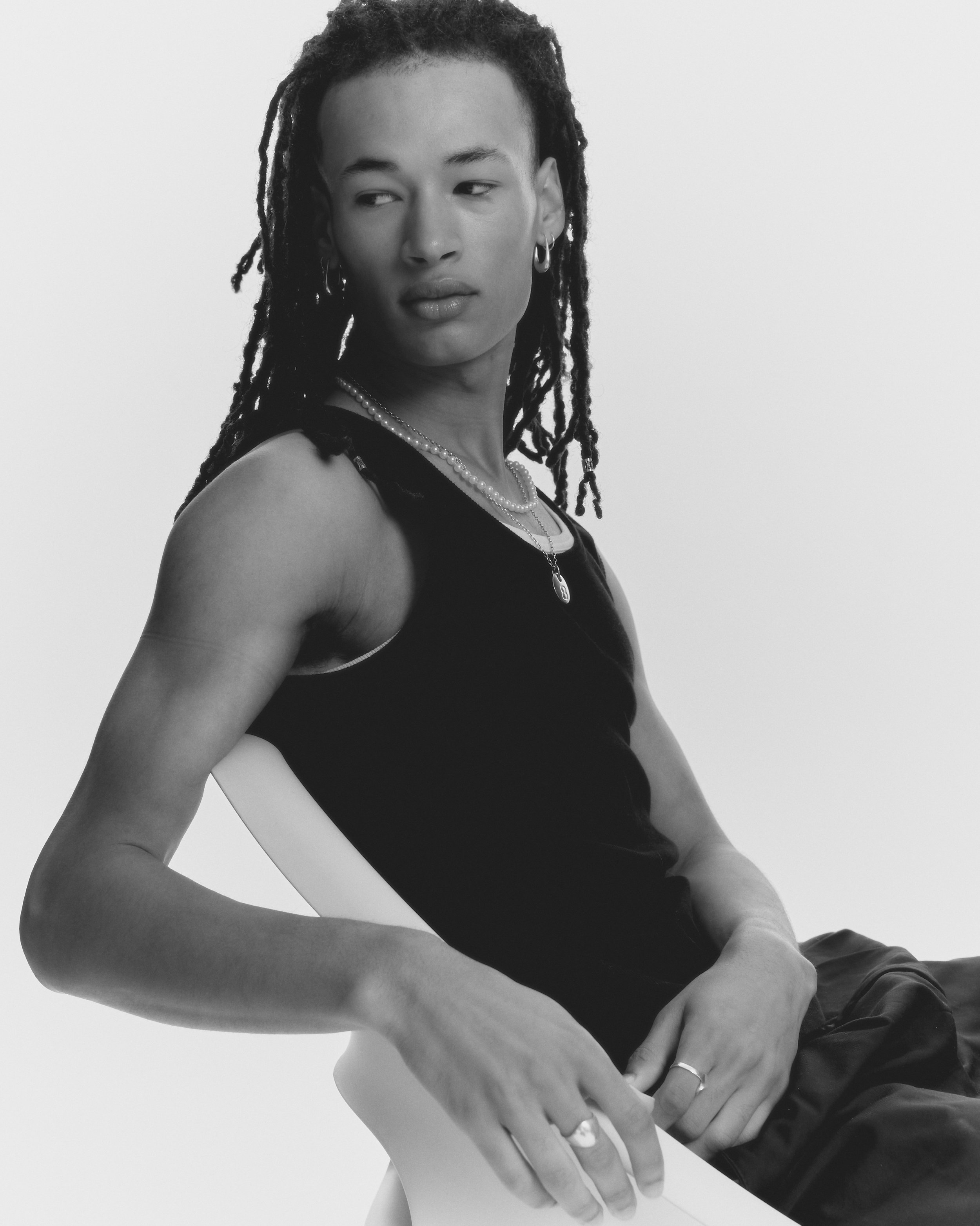 Young man with dreadlocks wearing a black tank top and silver jewellery, seated on a chair against a plain white background.