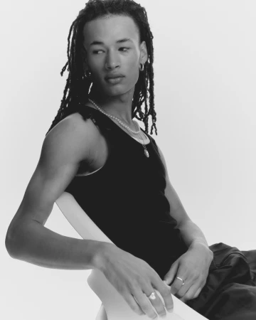 Young man with dreadlocks wearing a black tank top and silver jewellery, seated on a chair against a plain white background.