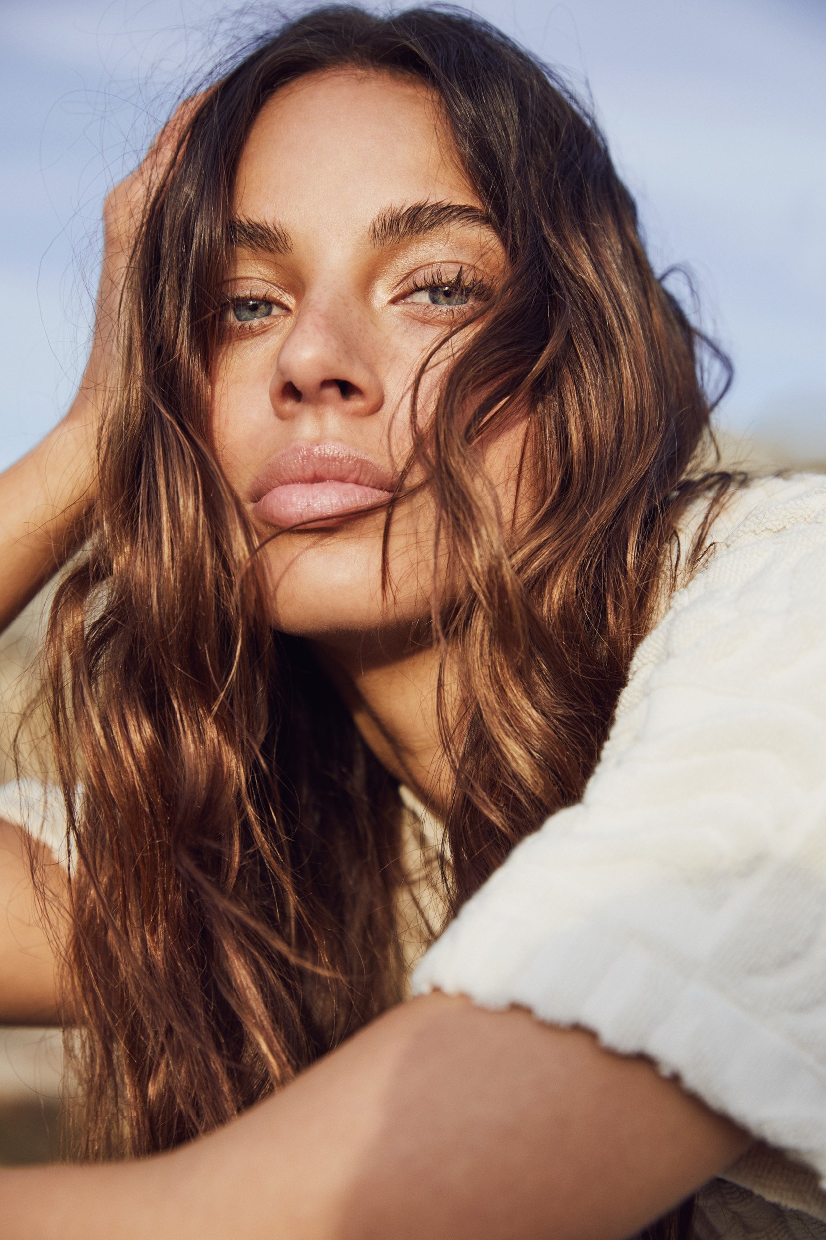 Close-up of an adult female model with long, wavy brown hair, fair skin, and blue eyes looking directly at the camera with a neutral expression.