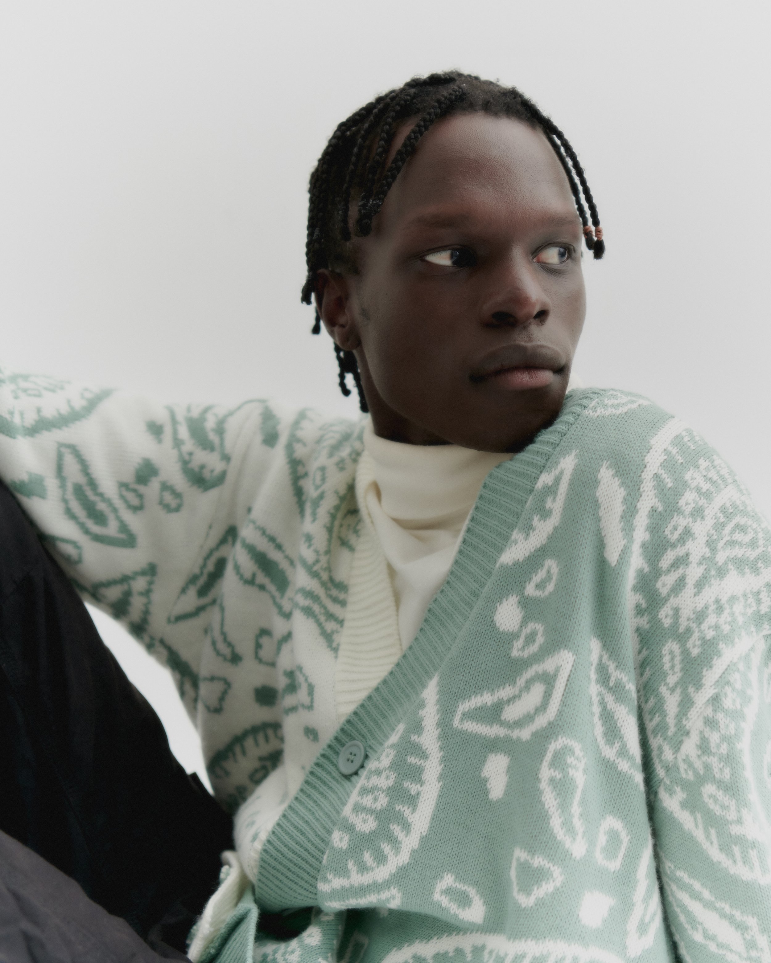 Portrait of an adult Black male in a patterned cardigan and turtleneck against a white background in a fashion forward style.