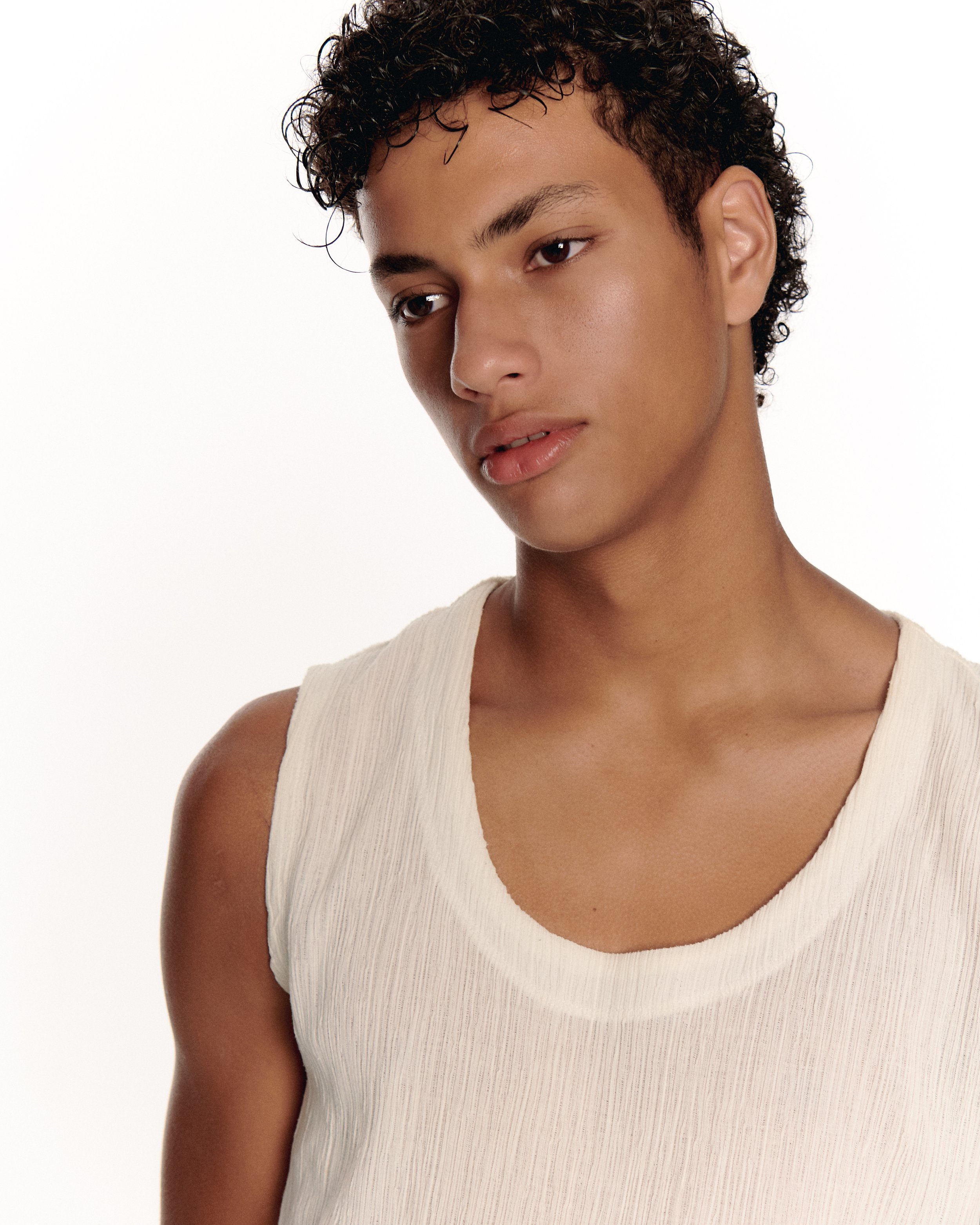 Close-up portrait of a young man with dark curly hair wearing a cream sleeveless top against a white background