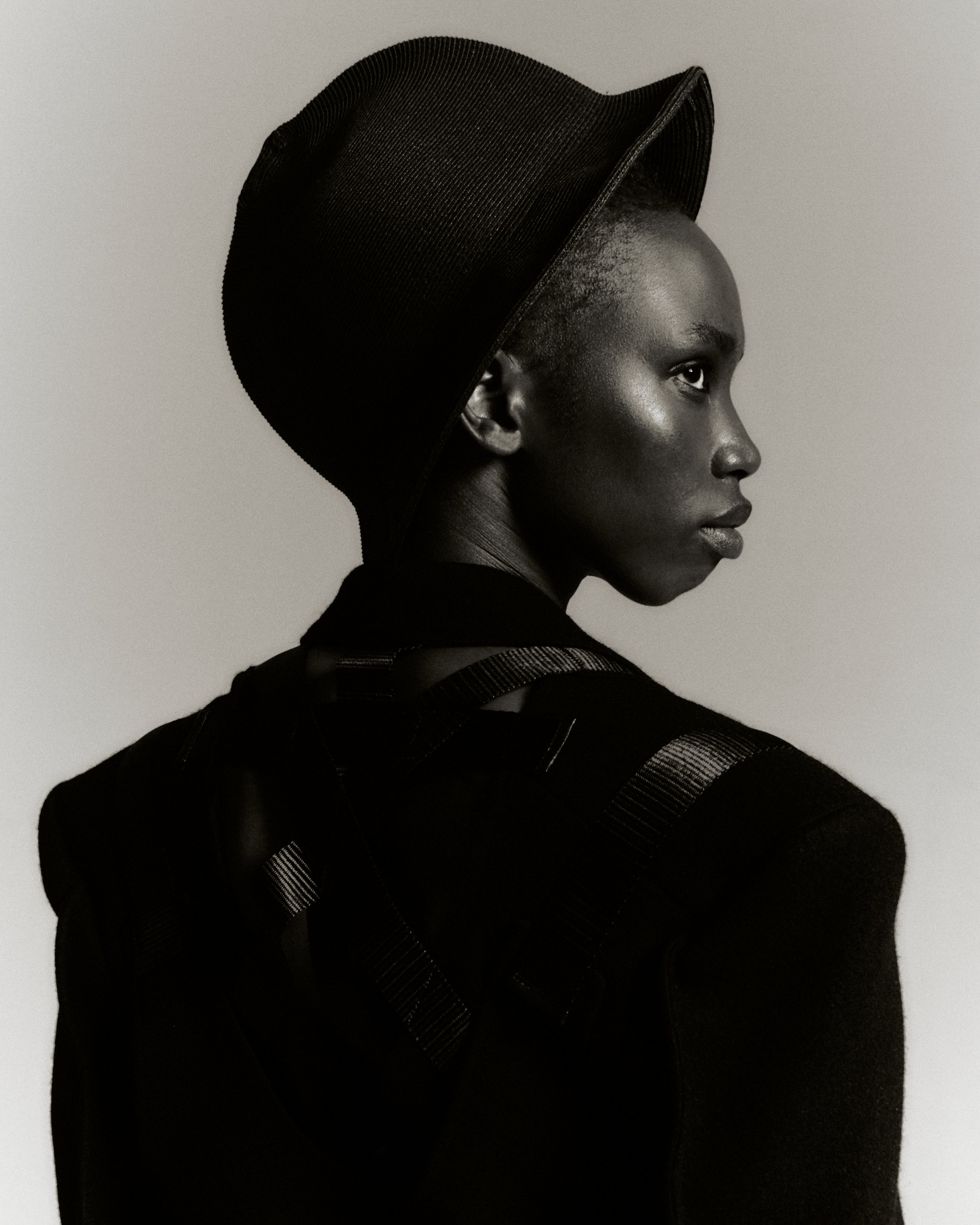 Monochrome portrait of an African female in a black structured hat and textured jacket, glancing to the right in a studio setting.