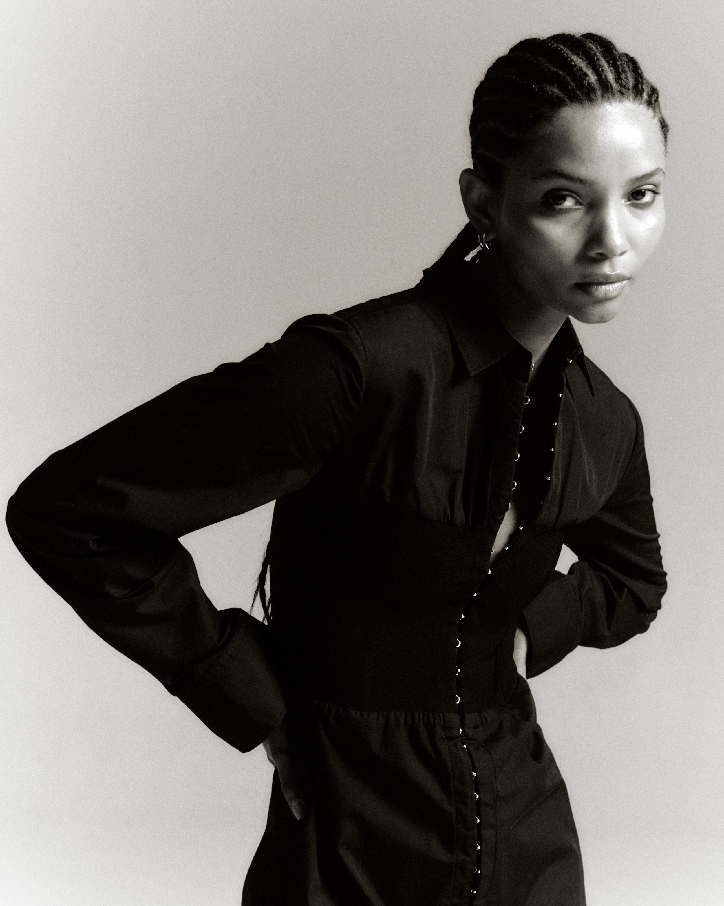 A stunning portrait of a Black woman with braided hair, wearing a stylish black shirt and corset, posing against a light background.