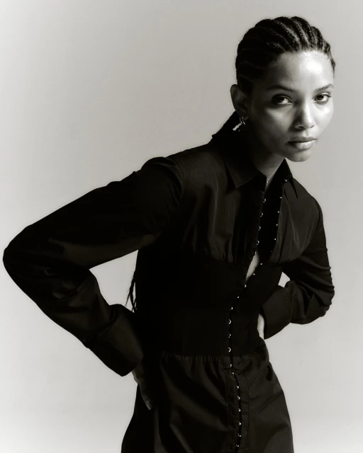 A stunning portrait of a Black woman with braided hair, wearing a stylish black shirt and corset, posing against a light background.