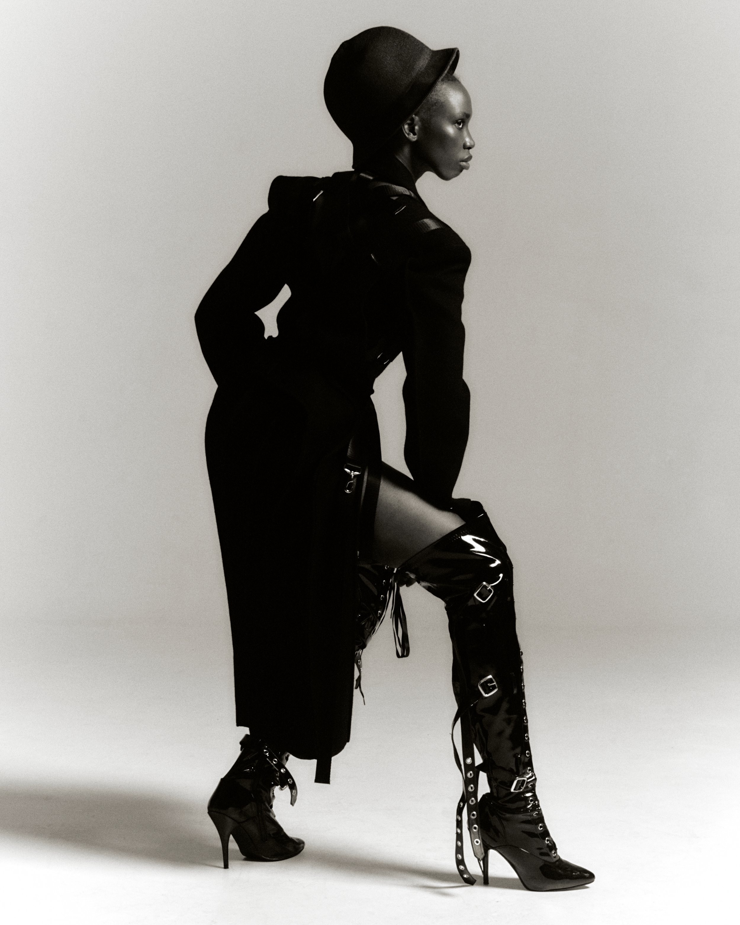 A striking monochrome fashion portrait of a Black woman in a black ensemble, featuring a structured coat, hat, and thigh-high buckled boots.