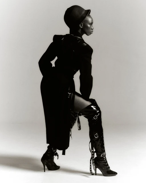 A striking monochrome fashion portrait of a Black woman in a black ensemble, featuring a structured coat, hat, and thigh-high buckled boots.