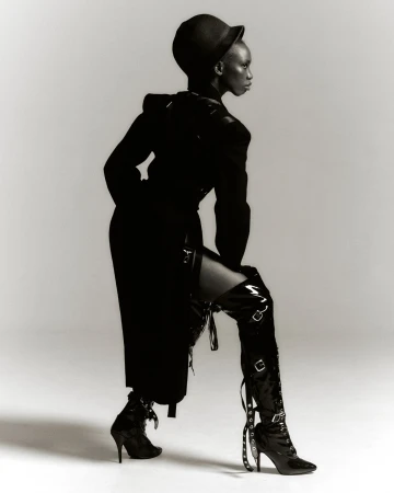 A striking monochrome fashion portrait of a Black woman in a black ensemble, featuring a structured coat, hat, and thigh-high buckled boots.