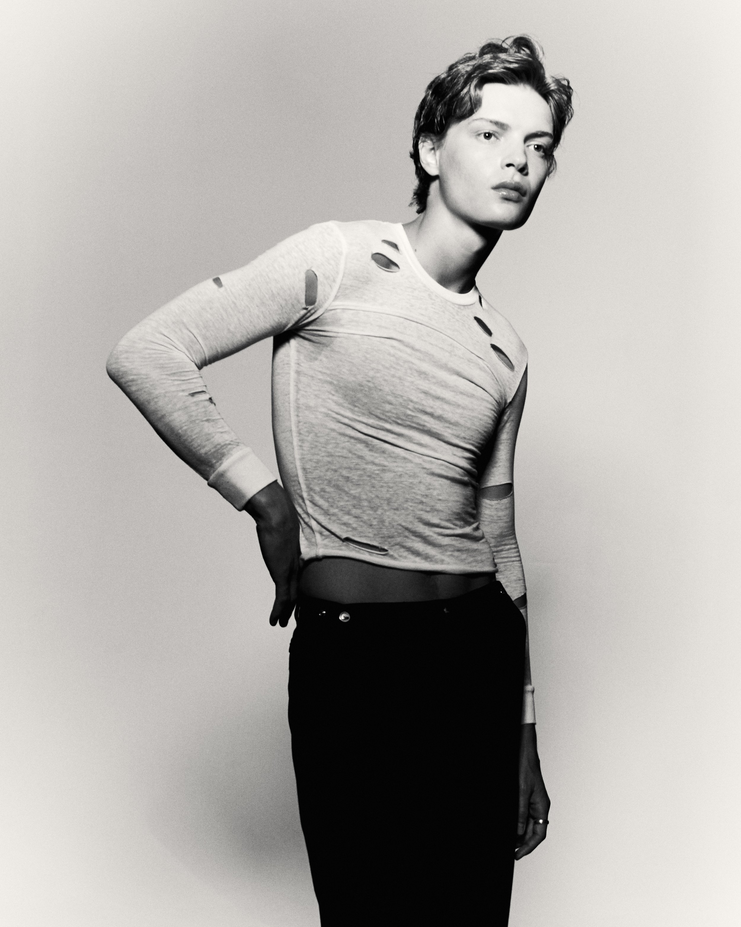 Monochrome portrait of a slender adult woman posing in a studio wearing a white long-sleeve top and dark bottoms against a solid background.