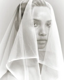 Close-up portrait of a woman wearing a sheer white veil over her head and face against a neutral background.
