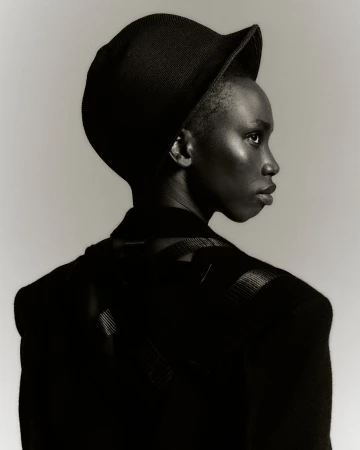 Black woman in profile wearing a dark cap and jacket against a neutral grey background