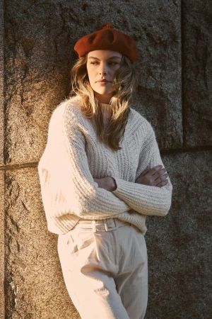 Woman in white knit sweater and rust beret leaning against stone wall with arms crossed outdoors in Sydney.