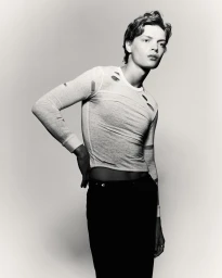Monochrome portrait of a young male model with styled hair wearing a distressed long-sleeve top against a light gradient background.