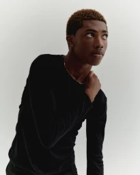 Black man in a black long-sleeve shirt with a gold chain necklace posing against a plain light background.