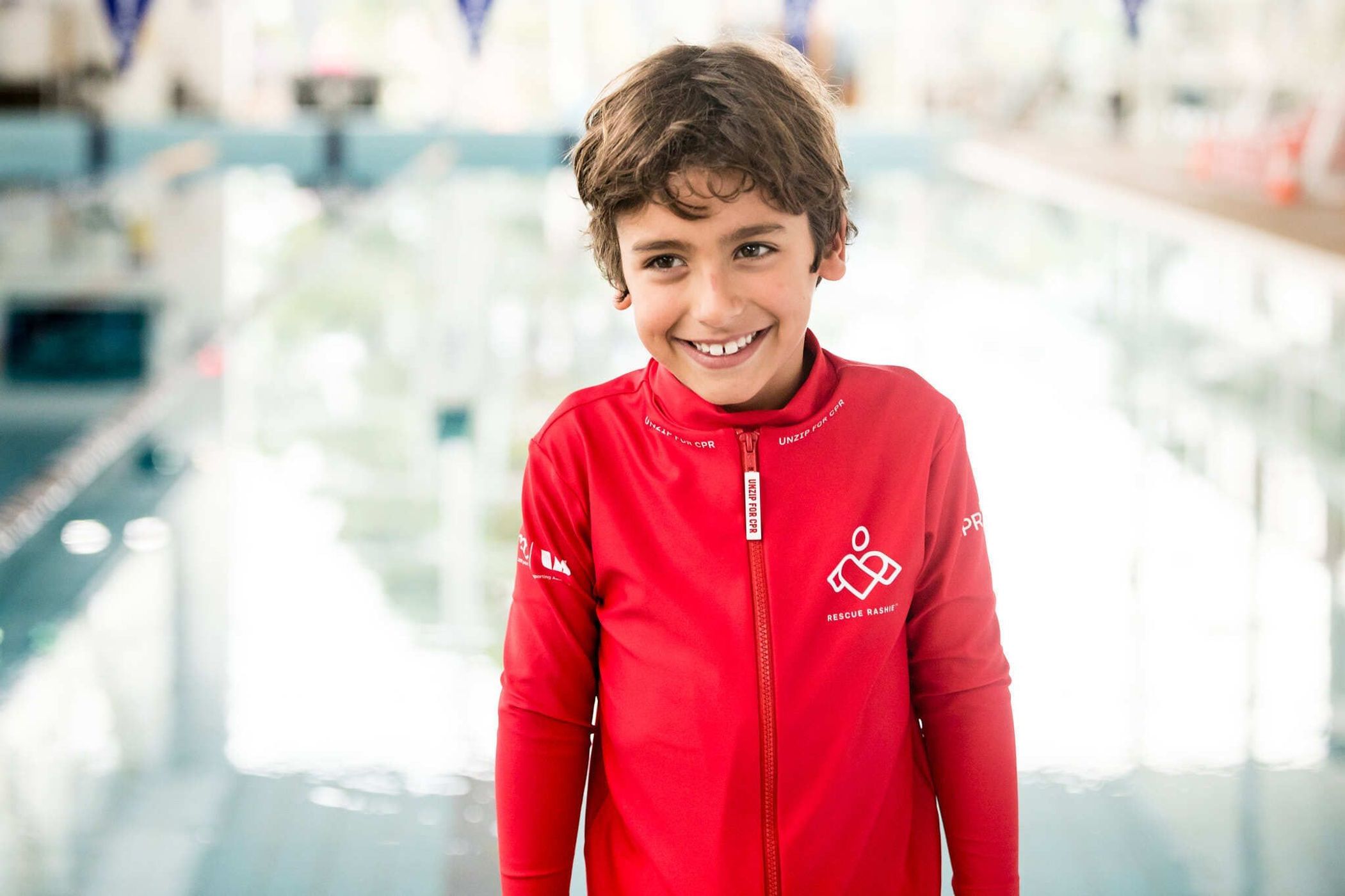 Smiling boy in a red jacket stands indoors near a swimming pool with a blurred background, portrait, eye-level angle.