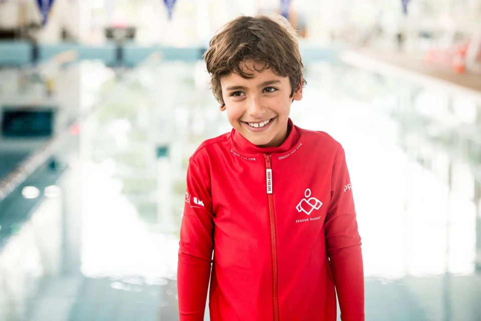 Smiling boy in a red jacket stands indoors near a swimming pool with a blurred background, portrait, eye-level angle.