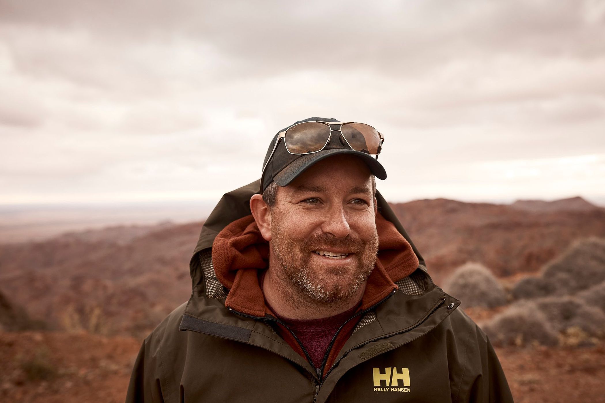 Close-up of a smiling adult male wearing sunglasses on his hat and an olive green jacket, with a desert landscape in the background.