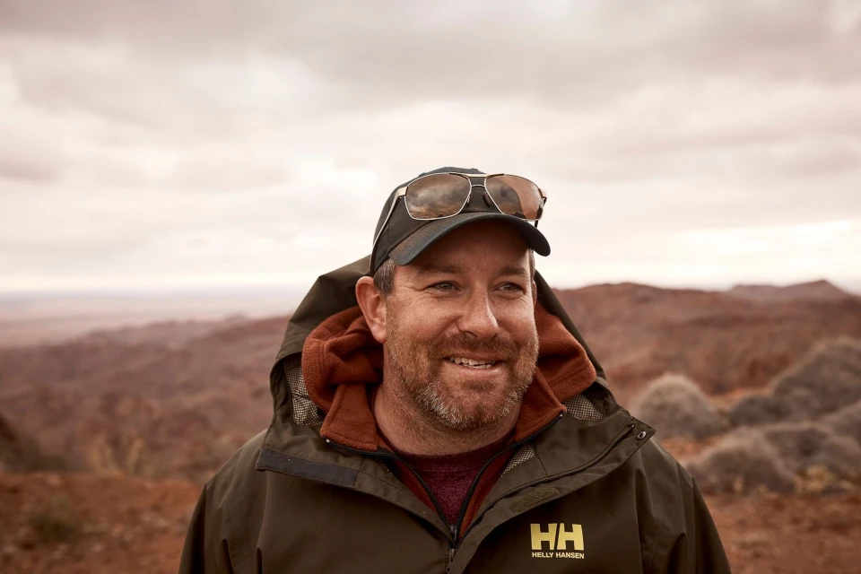Close-up of a smiling adult male wearing sunglasses on his hat and an olive green jacket, with a desert landscape in the background.