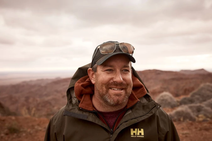 Close-up of a smiling adult male wearing sunglasses on his hat and an olive green jacket, with a desert landscape in the background.