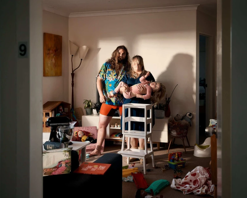 Indoor family portrait of a man, woman, and child in a messy living room with toys and furniture, basking in warm, natural light.