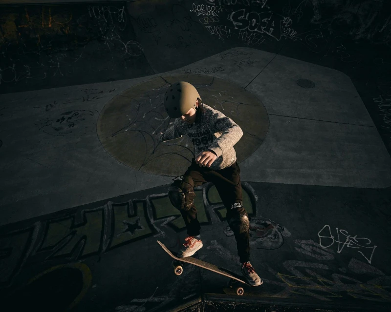 Youthful skateboarder in helmet and pads mid-air at a skate park performing an ollie, with graffiti-covered concrete walls.