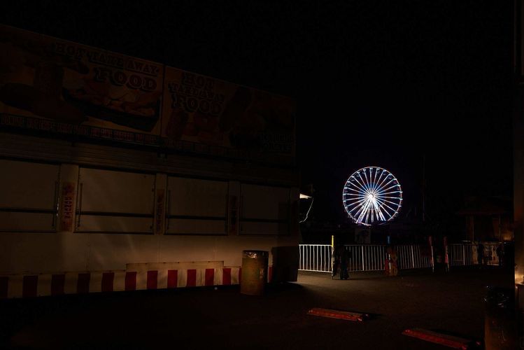 Night shot of a lit Ferris wheel in the distance, viewed from a dark street near an enclosed white vendor trailer.