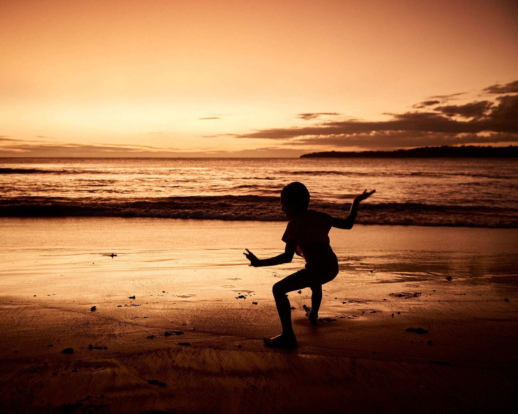 Child silhouette on a beach at sunset. The warm light casts shadows on the sand with ocean and horizon in the background.