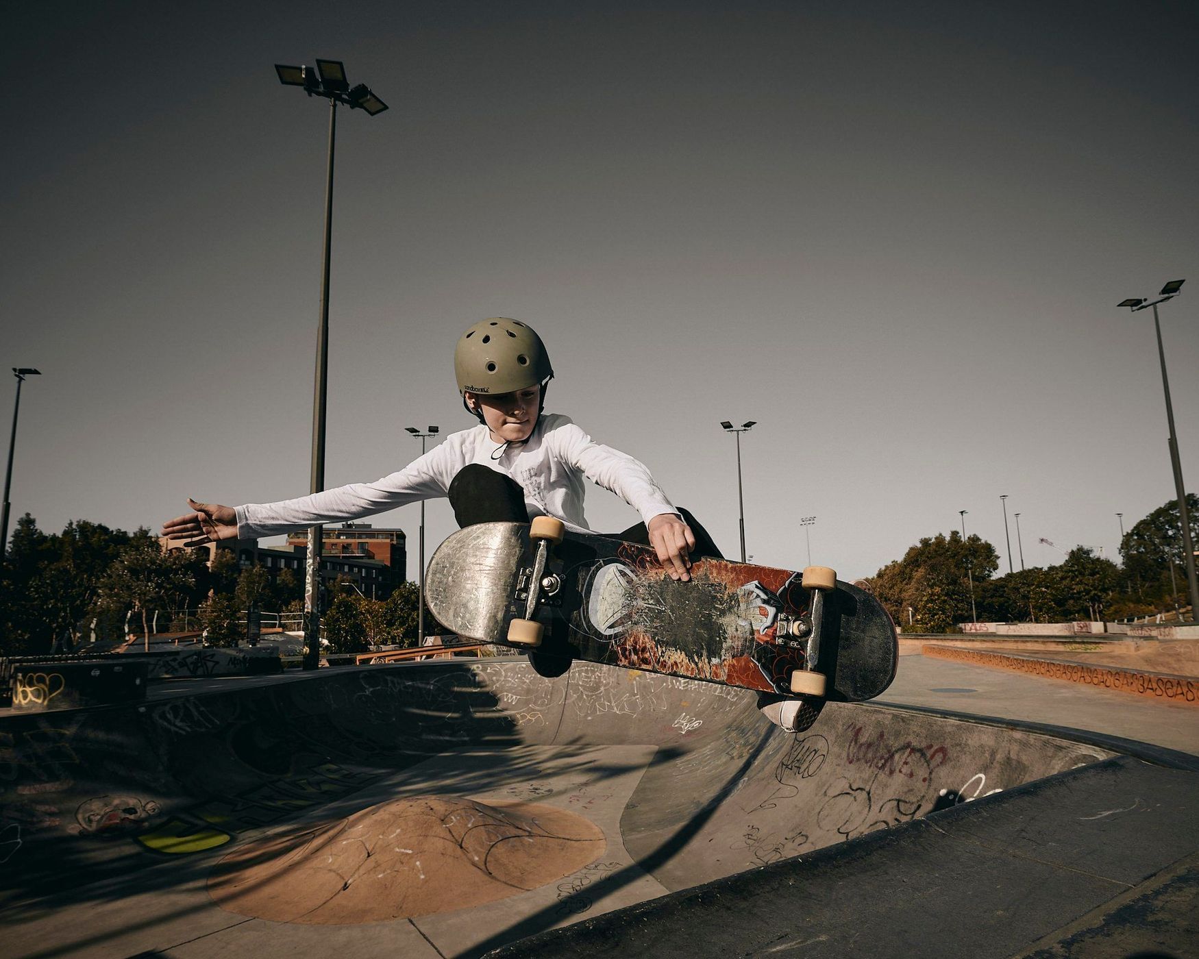 A youth wearing a helmet soars on a skateboard over a concrete ramp in an outdoor skatepark on a sunny day, performing tricks.