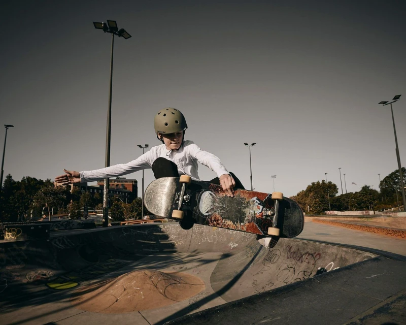 A youth wearing a helmet soars on a skateboard over a concrete ramp in an outdoor skatepark on a sunny day, performing tricks.