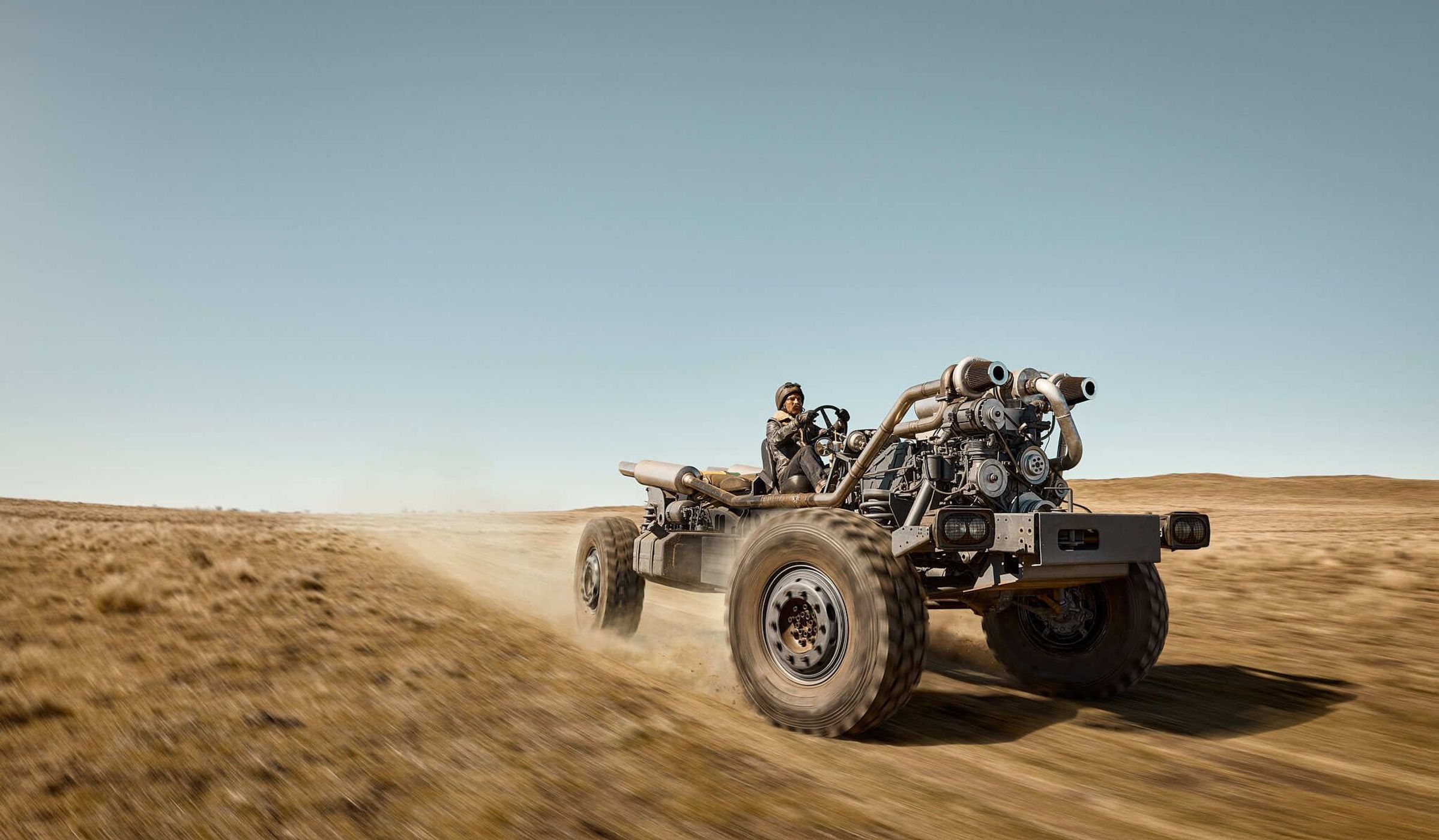 Modified vehicle with oversized tires in desert landscape, driven by a person in protective gear, against a clear blue sky backdrop.