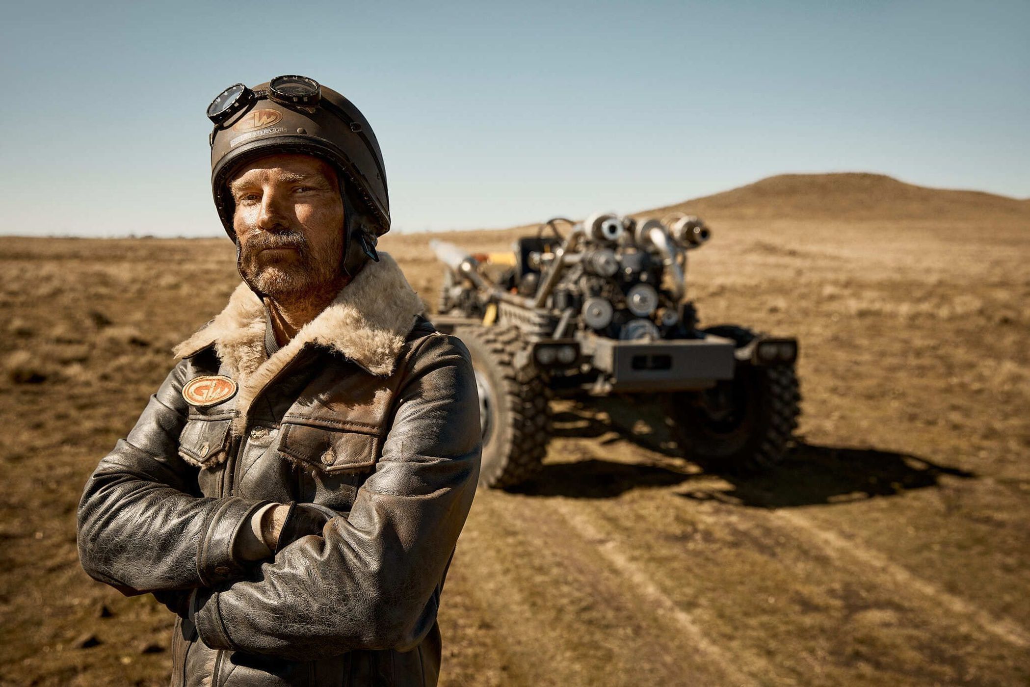Portrait of a rugged man in aviator helmet and leather jacket, arms crossed, next to an engine-powered vehicle in an arid landscape.