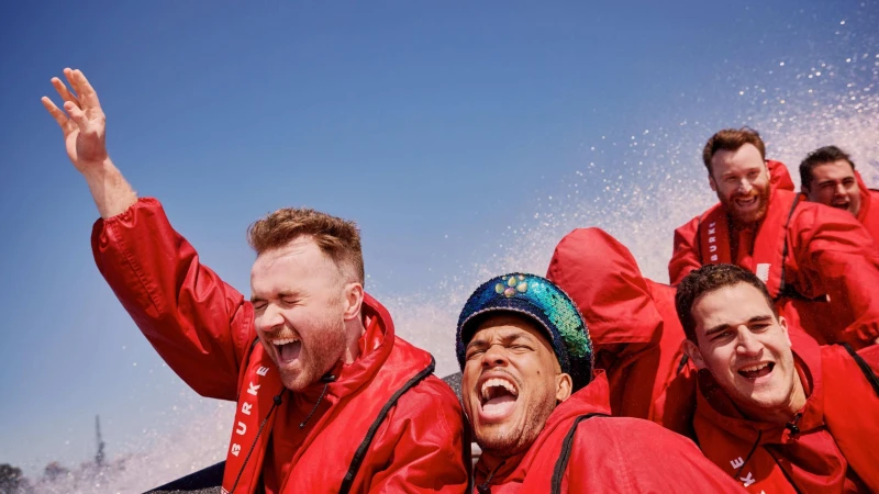 Enthusiastic group of six people in red jackets enjoying a boat ride on a sunny day with splashing water and a clear blue sky.