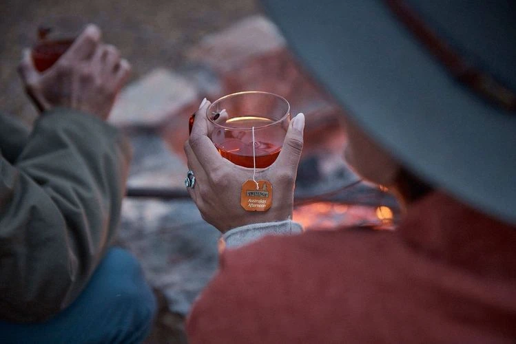 Two people drinking tea by the campfire. One person holds a steaming glass tea mug with a tea bag. Warm, cozy camping vibes.