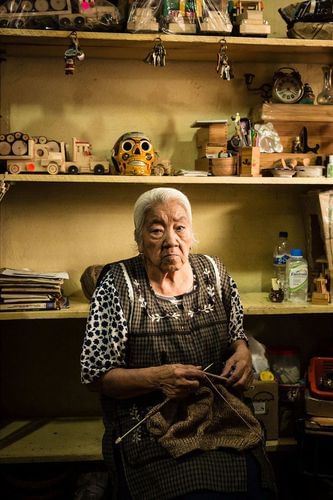Elderly woman sits indoors knitting in front of shelves stocked with wooden toys, a skull, and other objects in a dimly lit room.