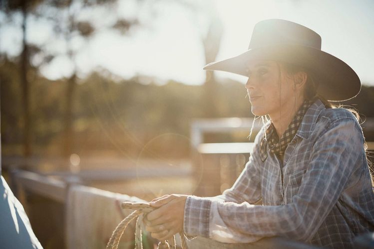 Adult female in cowboy hat and western wear holding rope at outdoor daytime location, with visible sunlight and rural background.