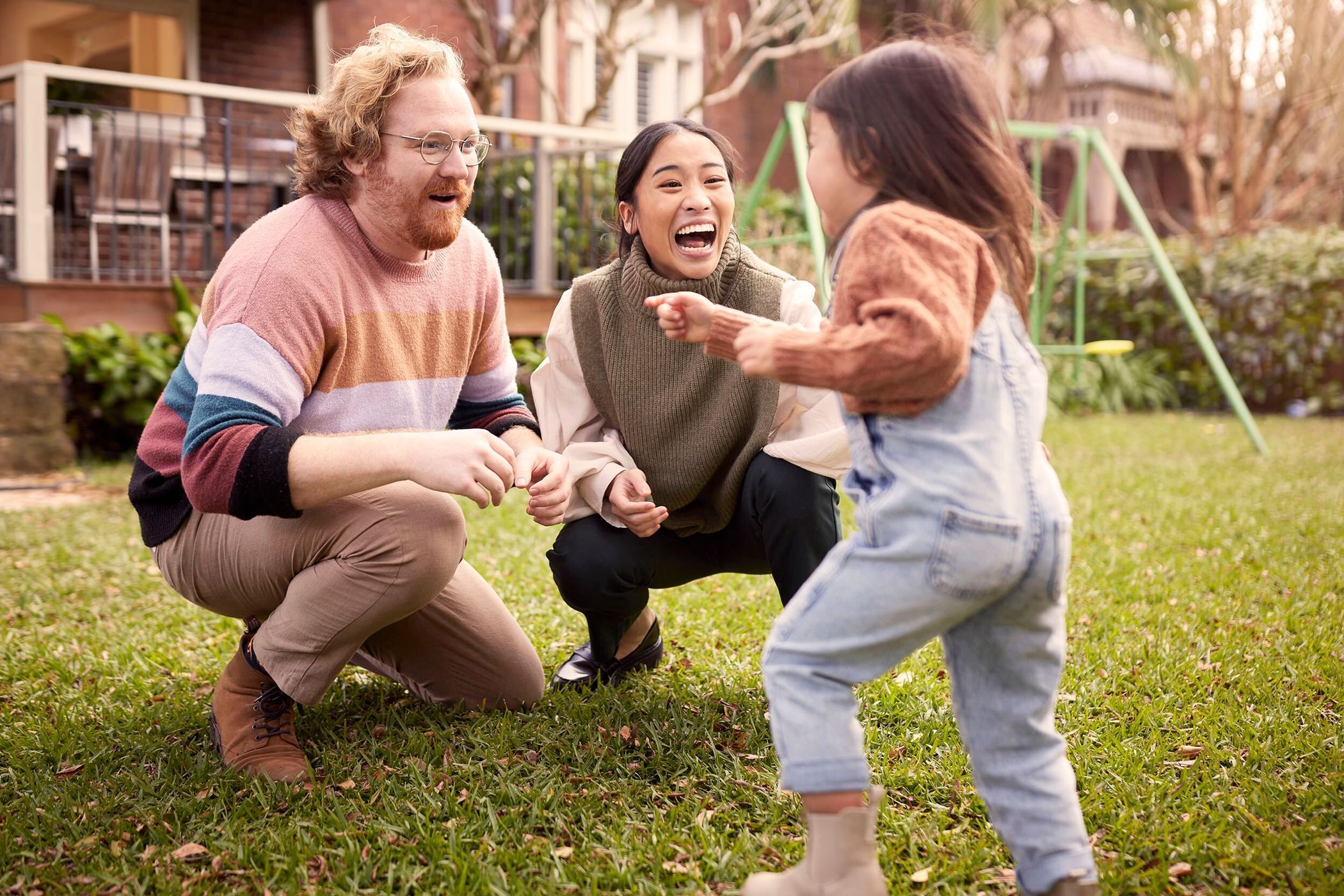 A family plays together in a grassy backyard. A father and mother watch a child, all smiling and happy, with a house in the background.