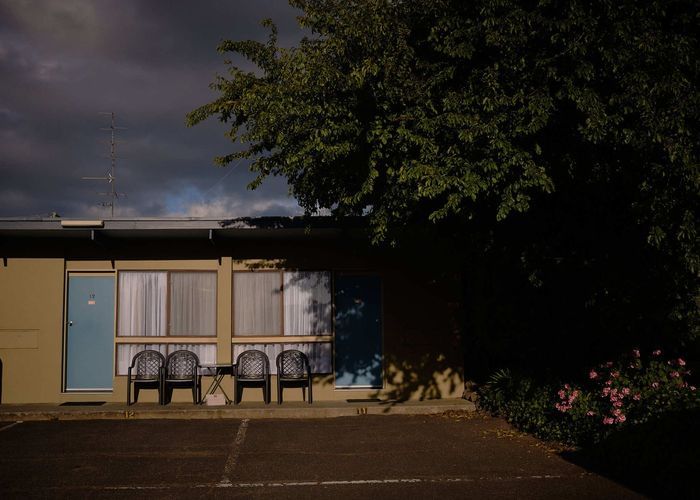 Motel facade with chairs outside rooms on a cloudy day. Dark tree looms over part of the building. Empty parking lot is in the foreground.