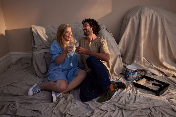 Smiling couple, an adult male and female, celebrate with clear glasses, sitting indoors on a paint-covered tarp, with paint can.