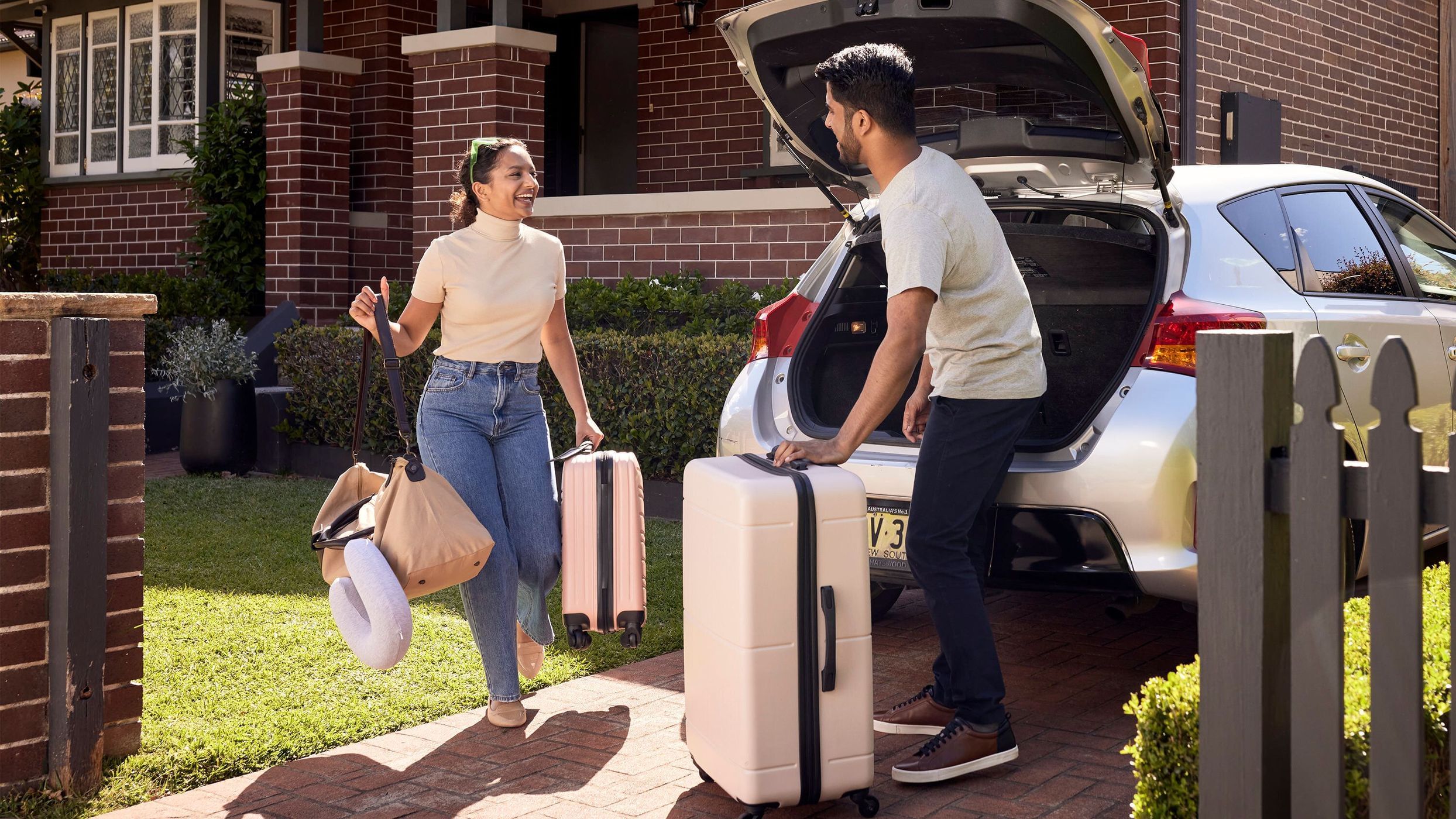 A couple loads luggage into the trunk of a silver hatchback car in front of a brick house with a green lawn and a wood fence.