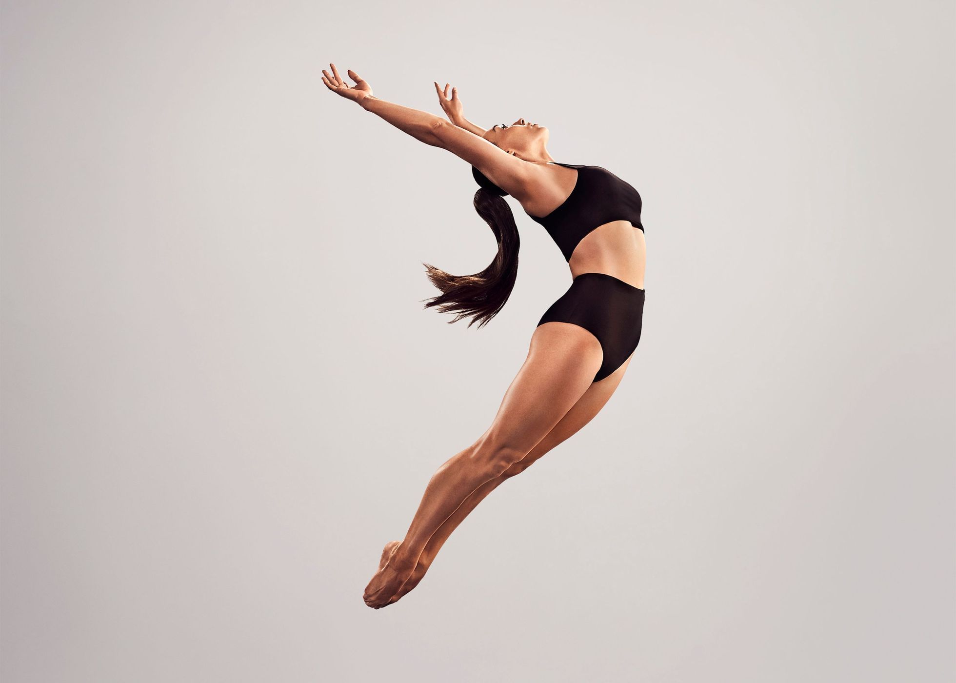 Woman in black athletic wear leaps mid-air against a neutral backdrop, arms raised, showcasing fitness and flexibility in a studio setting.