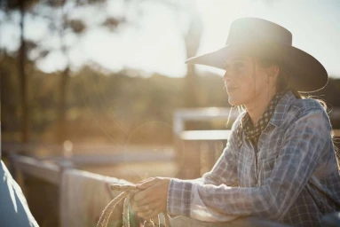 Woman in cowboy hat and plaid shirt leaning on a fence outdoors at sunset by photographer Klint Collier