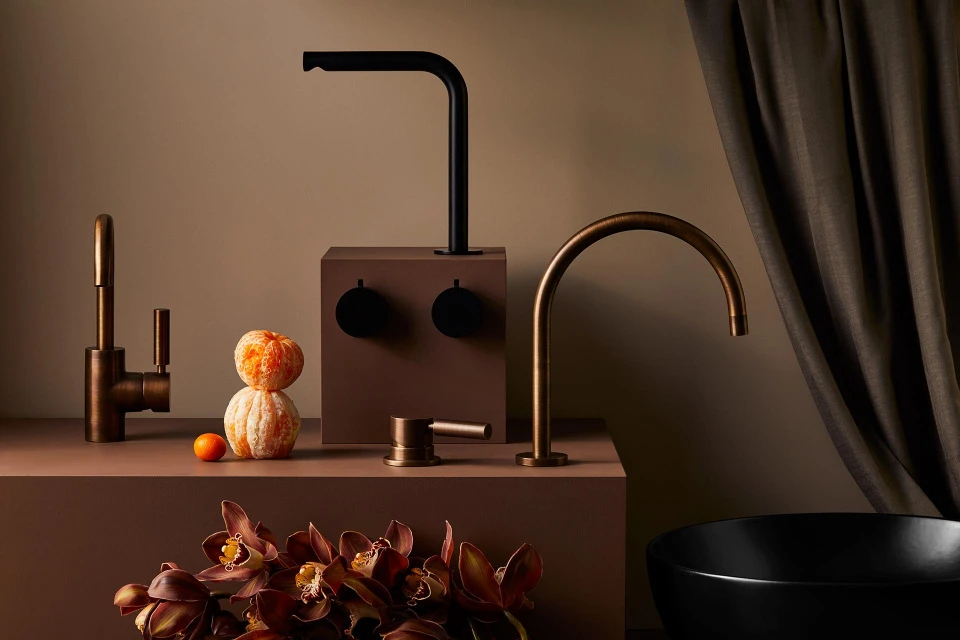Still life of modern black and bronze bathroom faucets arranged on brown platforms with decorative oranges and dark floral accents.