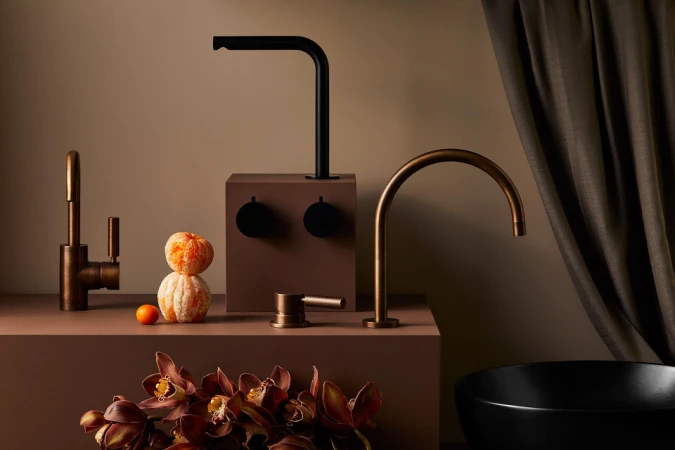 Still life of modern black and bronze bathroom faucets arranged on brown platforms with decorative oranges and dark floral accents.