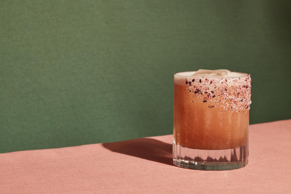 Cocktail in a rocks glass with salt and dark red powder around the rim on a pink table against a two-tone green background.
