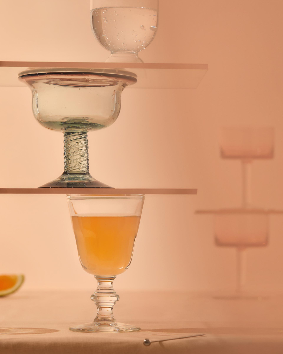 Stacked glass stemware on wooden planks displays cocktails and still water against a peach-colored backdrop with a cocktail spoon on table.