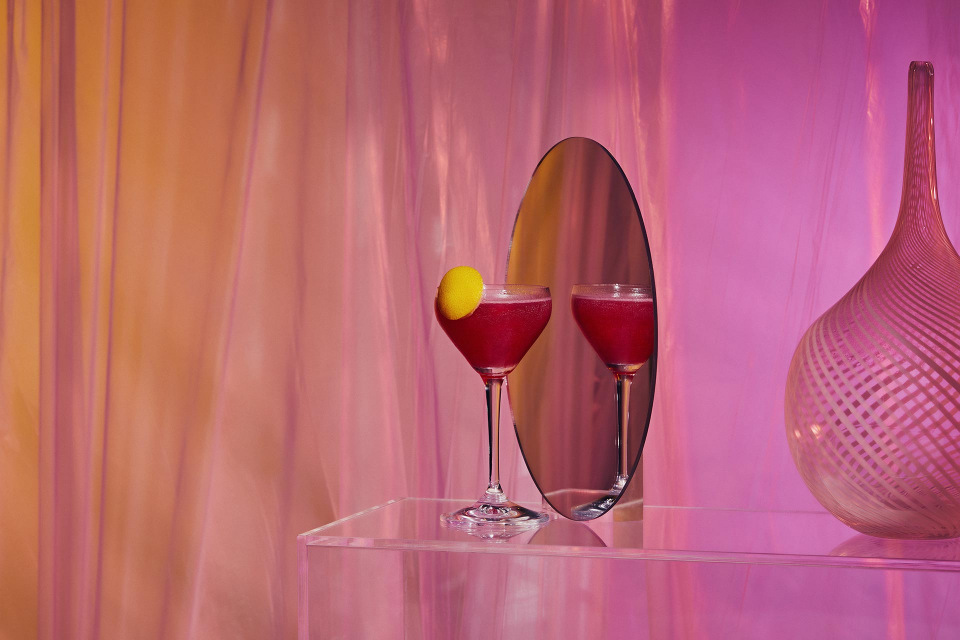 Two magenta-colored cocktails in glasses on a clear table next to a decorative vase in front of pink fabric backdrop.