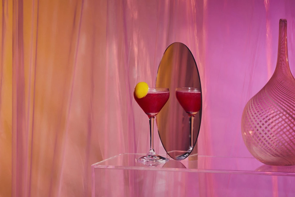 Two magenta-colored cocktails in glasses on a clear table next to a decorative vase in front of pink fabric backdrop. 