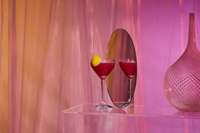 Two magenta-colored cocktails in glasses on a clear table next to a decorative vase in front of pink fabric backdrop. 